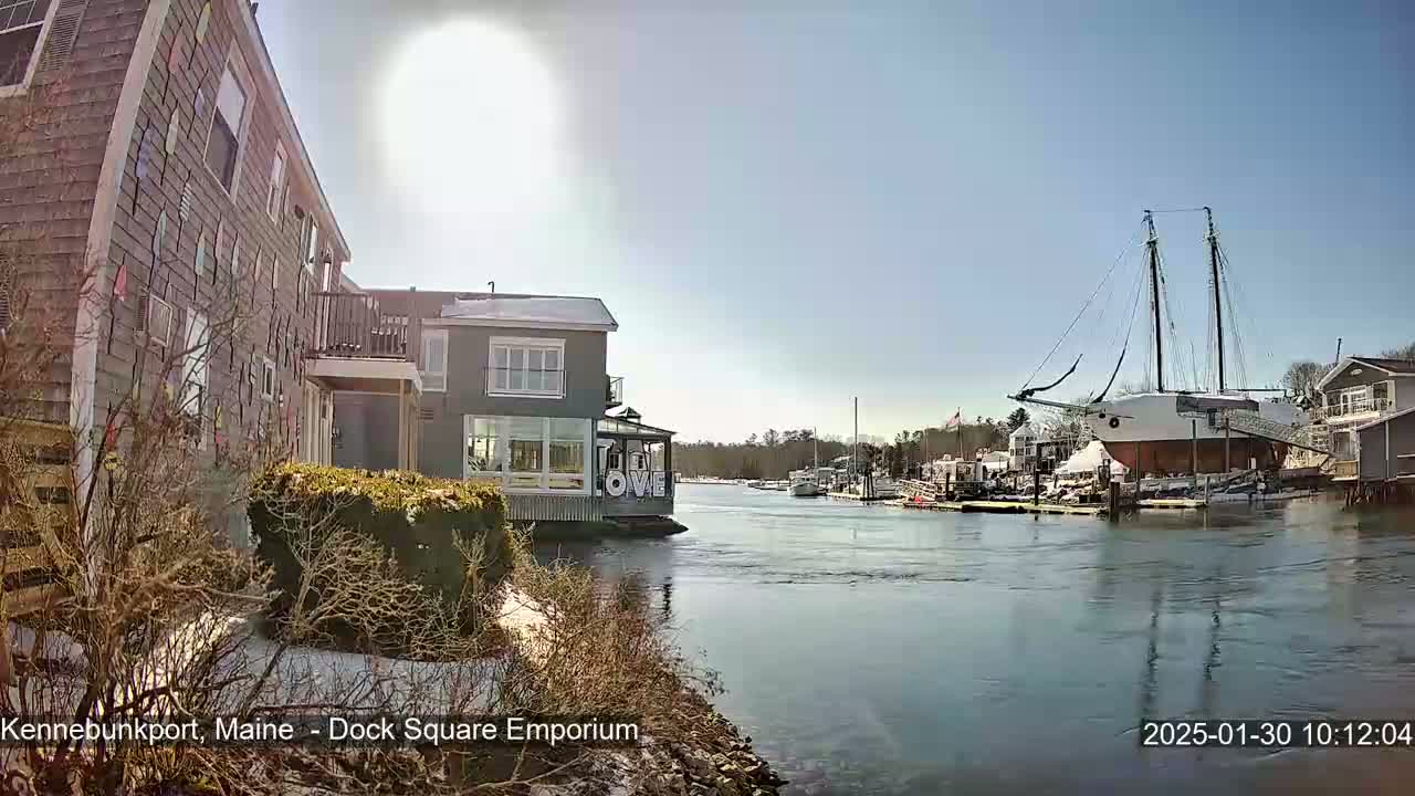 A sunny day reveals a waterfront scene with a large sailing ship docked near buildings, some with snow on the ground.