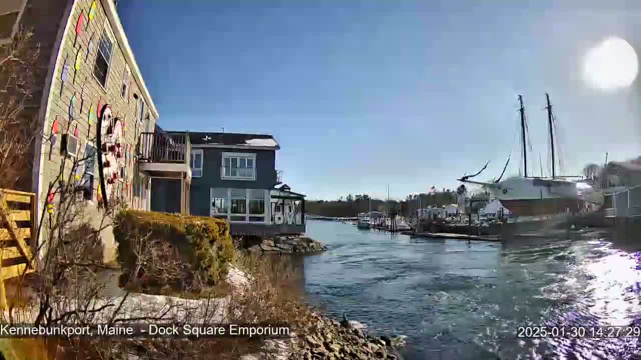 A colorful building sits beside a calm waterway on a sunny day, with a large sailboat docked nearby and other boats in the distance.