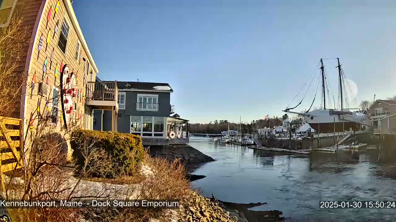 A waterfront scene on a sunny day shows a building decorated with colorful buoys, other buildings along a calm river, and a large sailing ship docked nearby.