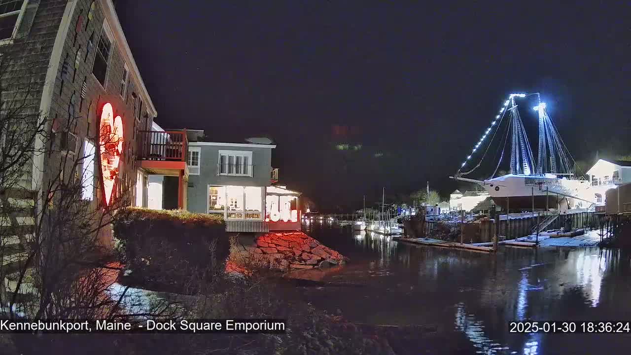 A nighttime waterfront scene shows a building with a large red heart-shaped light display next to a calm waterway, across which a large ship is illuminated, all lightly dusted with snow.