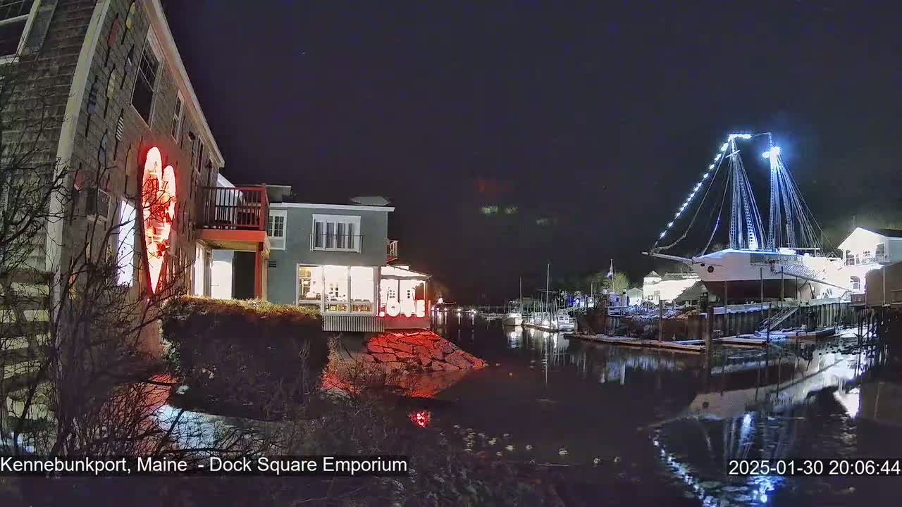 A nighttime scene shows a waterfront area with buildings lit up, a large sailboat docked, and a calm body of water reflecting the lights.