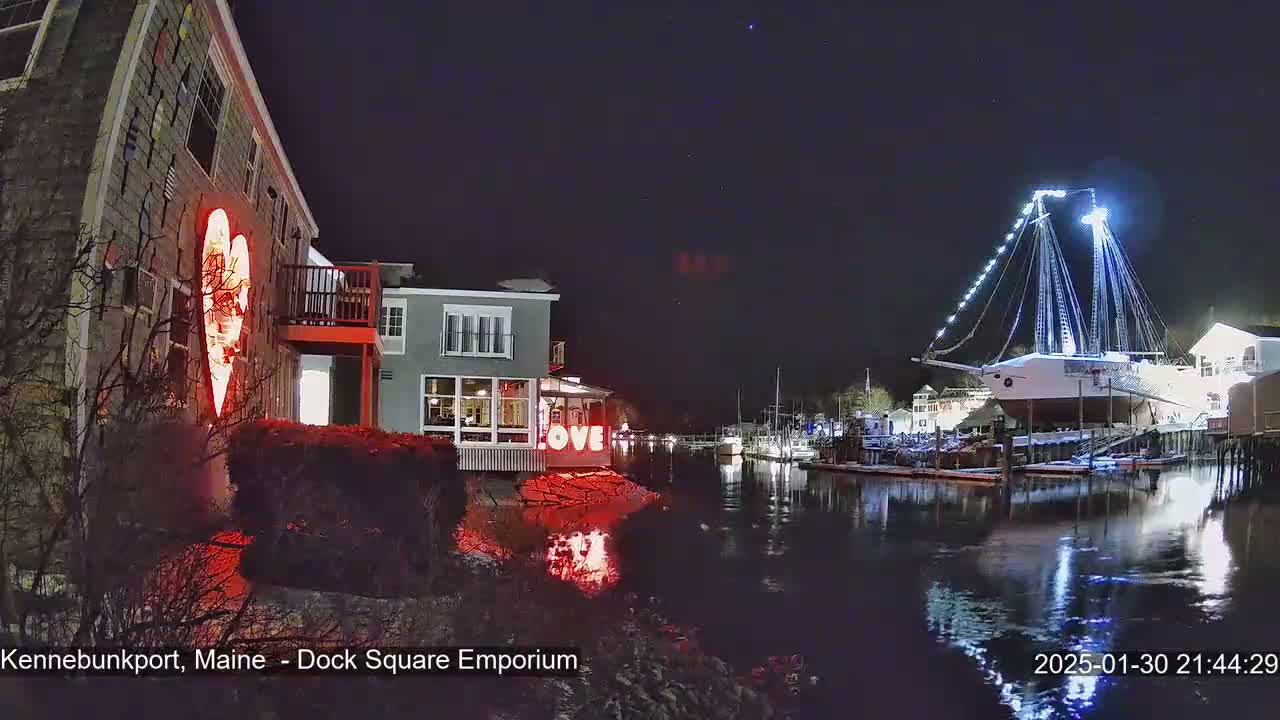 A nighttime waterfront scene shows a building with a large illuminated heart on its side, a  lit-up ship in the water, and other buildings along the shore under a clear, dark sky.