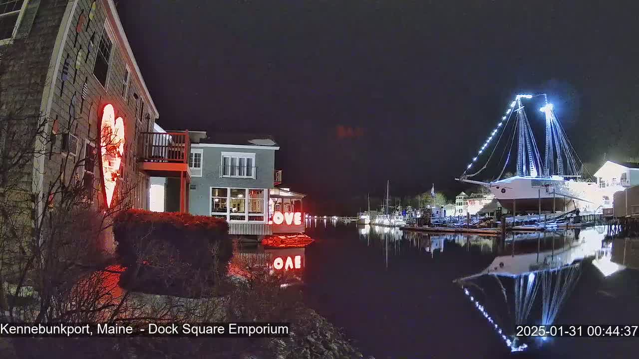 A nighttime harbor scene shows a building with a large illuminated heart, a waterfront building with "LOVE" displayed, and a large illuminated sailboat docked at the water's edge, all reflecting in the calm water.
