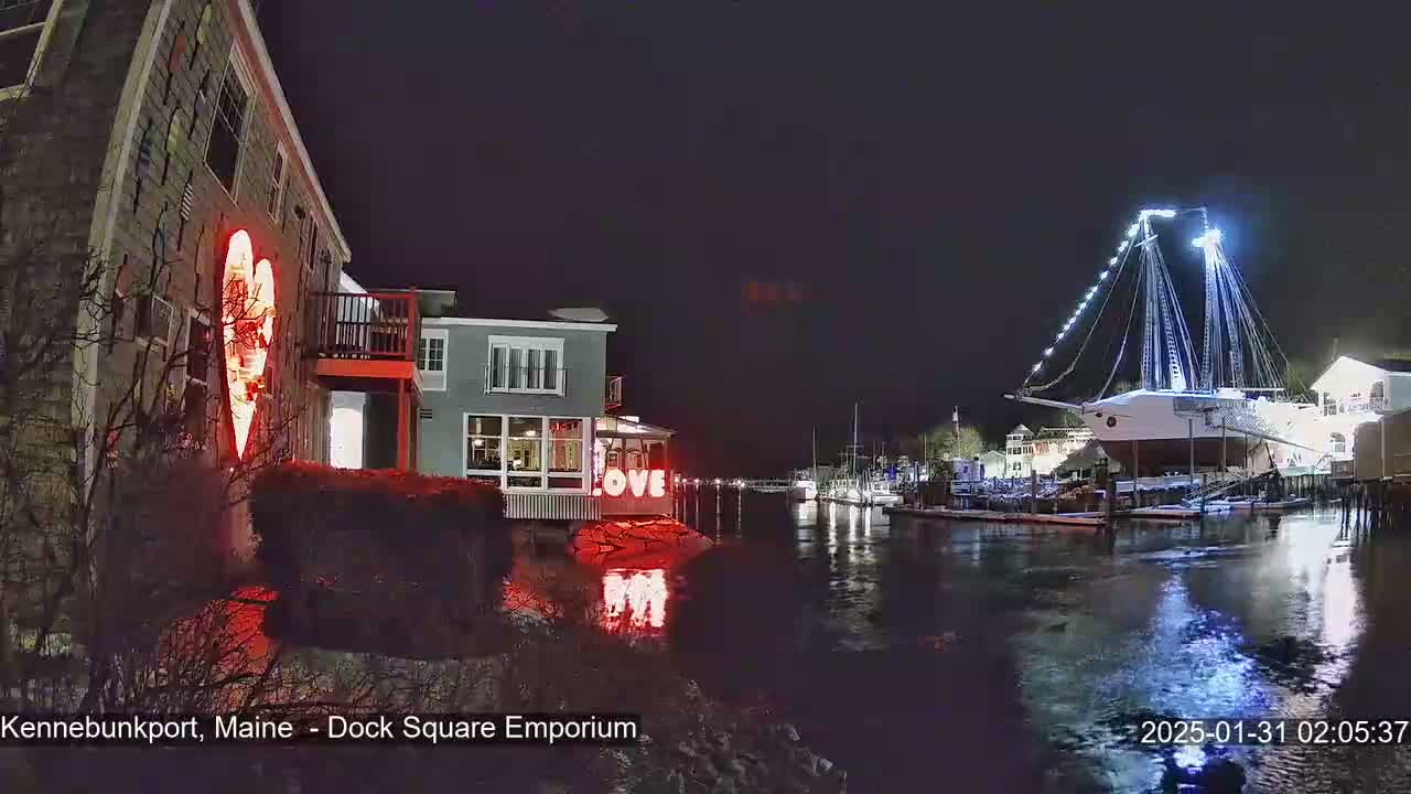 A nighttime waterfront scene shows a building with a large illuminated heart on its side, another building with illuminated "LOVE" letters, and a large sailboat illuminated with lights, all reflected in calm water.