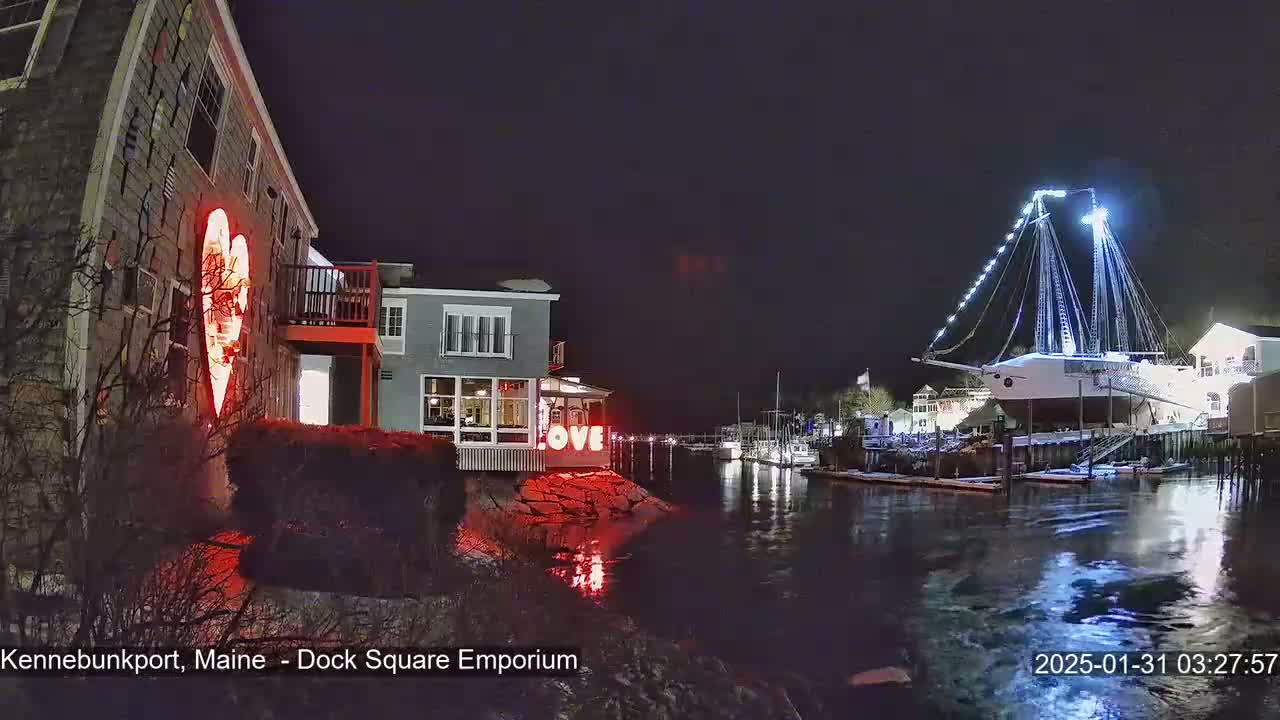 A nighttime waterfront scene shows a building with a red illuminated heart, another building with a "LOVE" sign, and a large ship illuminated with white lights docked in the water.