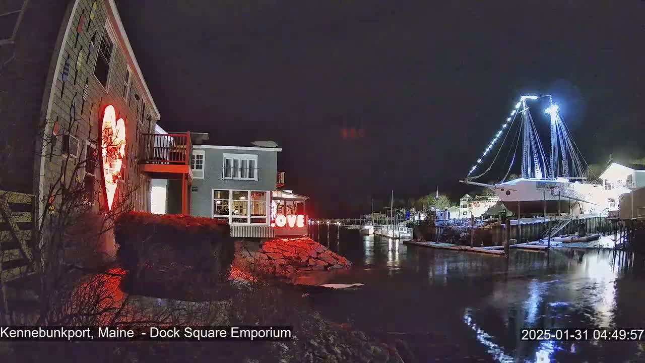 A nighttime scene shows a waterfront area with a building illuminated by a large red heart and the word "LOVE," and a large sailboat docked, brightly lit against a dark sky.