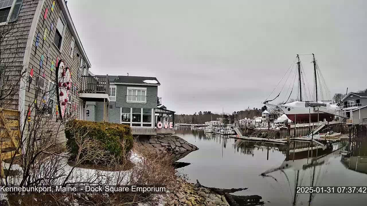 A waterfront scene on an overcast day shows buildings, a large sailboat on dry dock, and a calm waterway reflecting the surroundings.