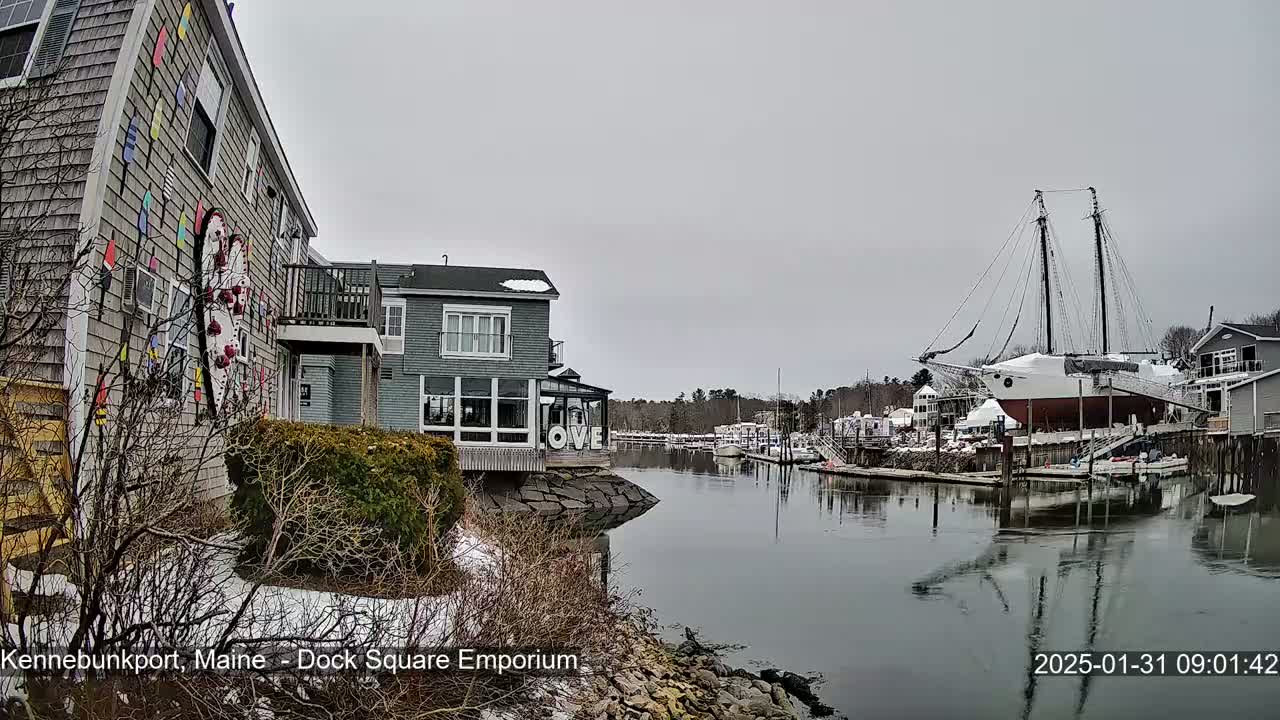 A harbor scene with snow on the ground and overcast skies shows a large sailboat docked near buildings with colorful decorations, and other boats in the background.
