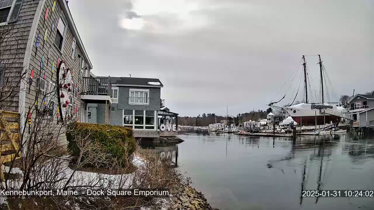 A waterfront scene on an overcast day shows a gray building with colorful artwork, other buildings, a large sailboat docked, and a calm waterway with snow on the ground.