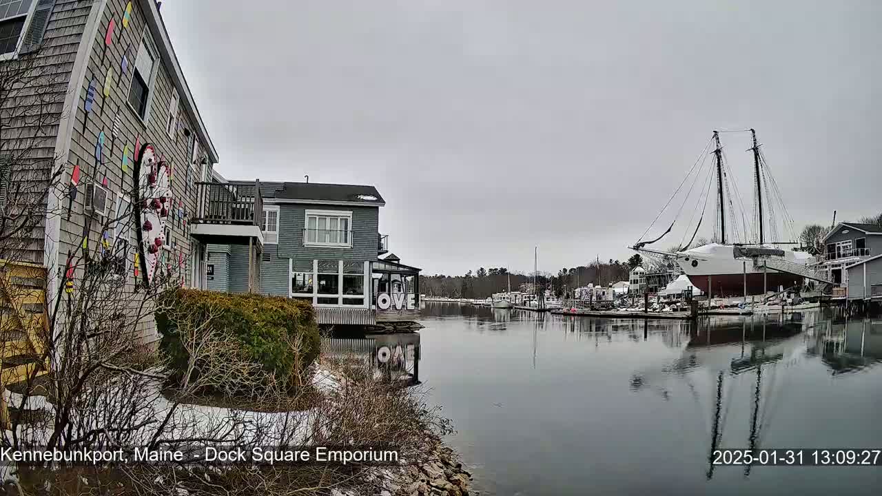 A calm harbor with a large sailboat docked near colorful buildings on a partly cloudy day with patches of snow on the ground.