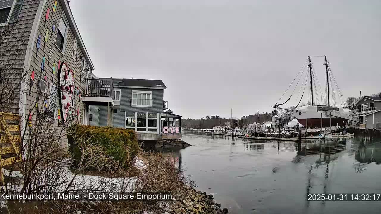 A gray, overcast day shows a waterfront scene with a large sailboat docked near buildings, some of which have snow on the ground, and a calm harbor.