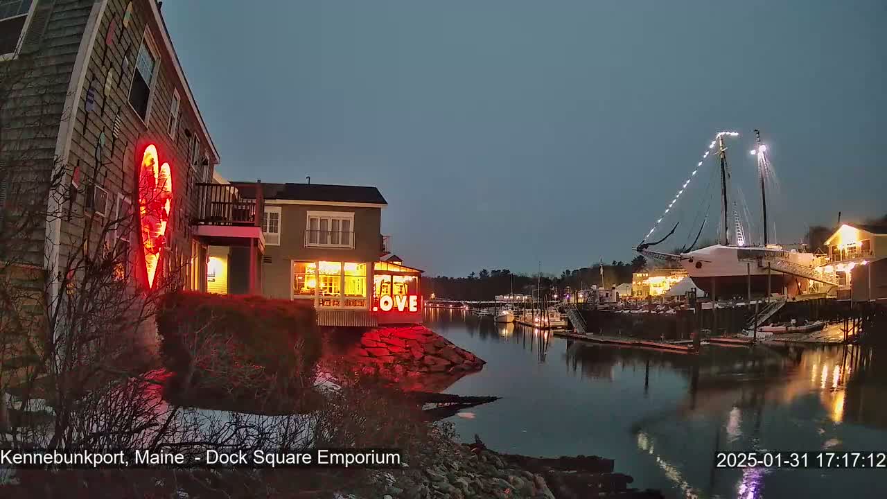 A nighttime waterfront scene shows a building with a large illuminated heart, other lit buildings, a sailboat docked, and a calm body of water with some snow on the ground.