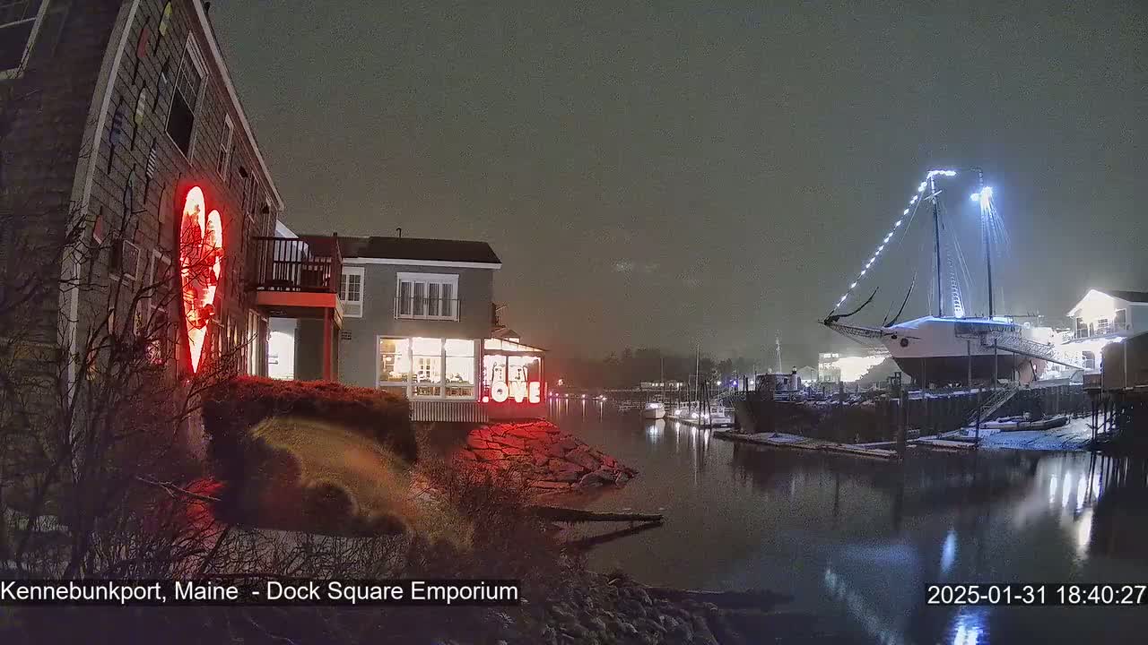 A nighttime scene of a snow-dusted harbor with a large sailboat docked near illuminated buildings, one of which displays a large red heart.