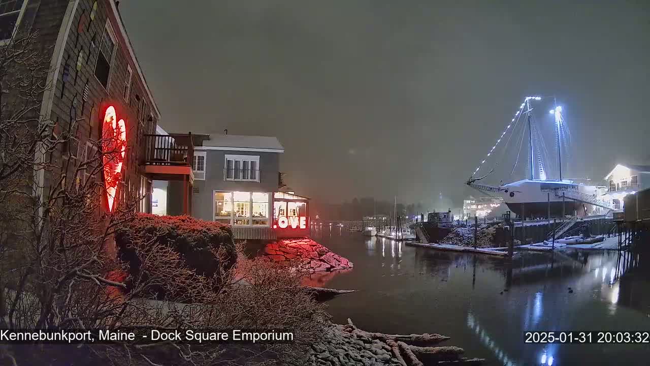 A snow-covered waterfront scene at night shows a building with a large illuminated heart, another building with a "LOVE" sign, and a large sailboat docked, all illuminated under a dark, snowy sky.
