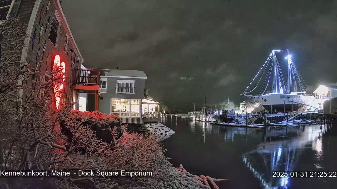 A snow-covered waterfront scene at night shows a large sailboat illuminated with lights, docked near buildings with a red heart-shaped light on one.