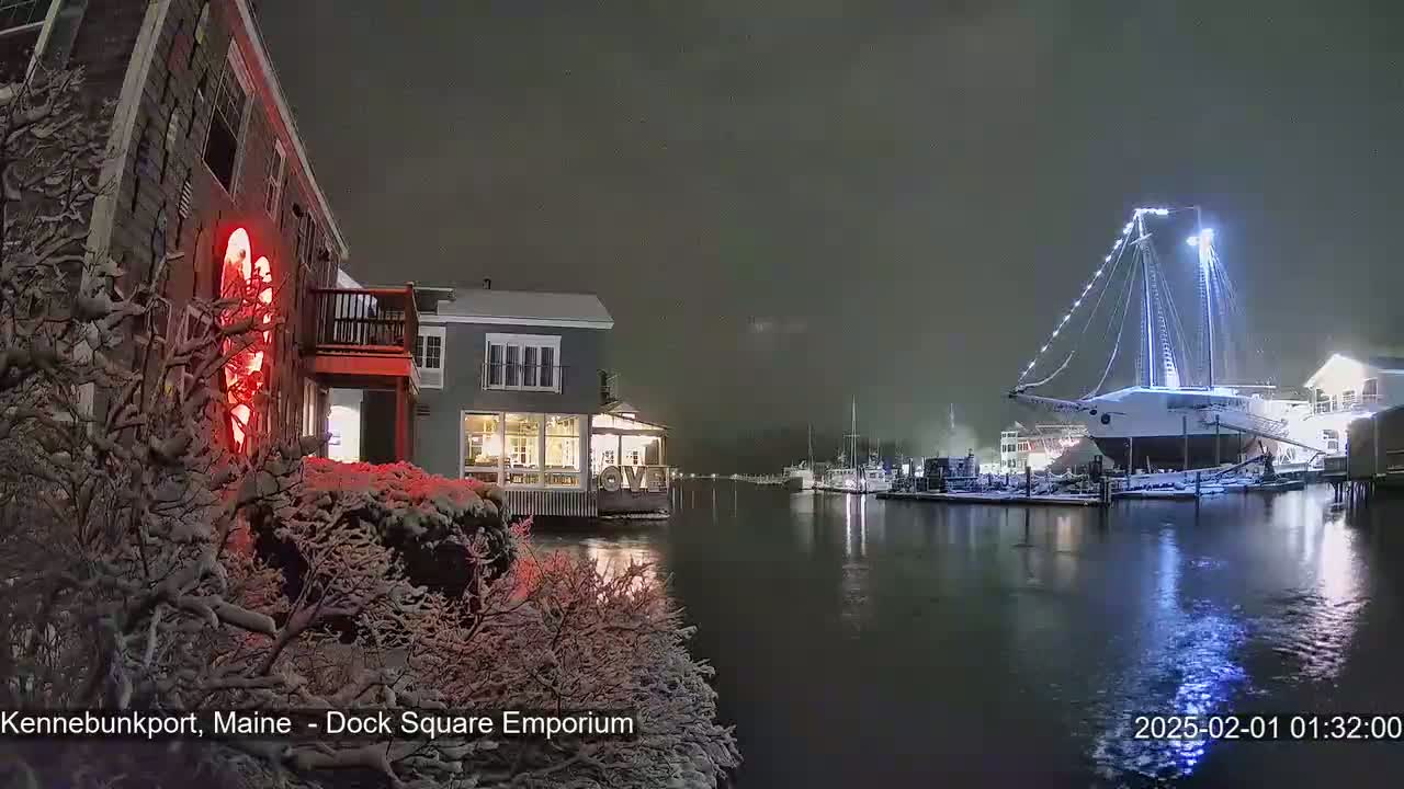 Snow-covered buildings and a large, illuminated sailboat are visible at night near a body of water.