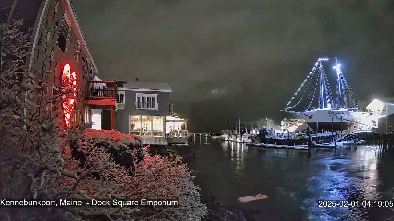 A snow-covered waterfront scene at night shows a large, illuminated sailboat docked near lit buildings.