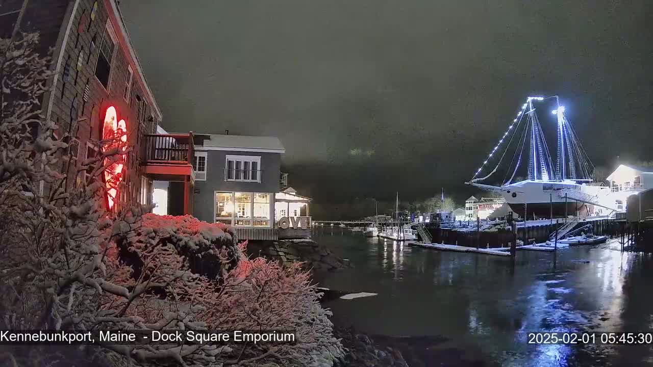 A snow-covered waterfront scene at night shows a large sailboat docked near buildings with lights on, under a dark, cloudy sky.