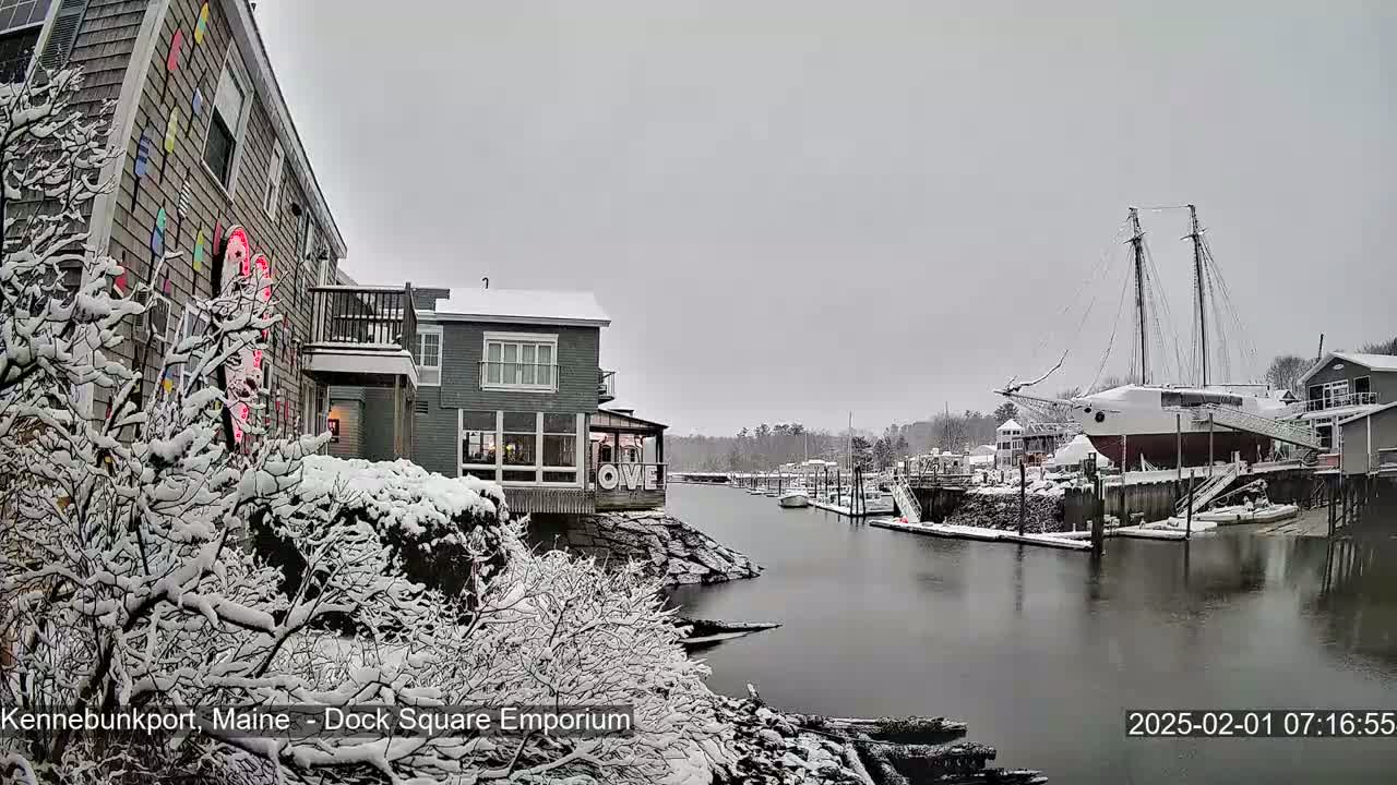 Snow-covered buildings and a docked sailboat are seen next to a waterway under an overcast sky.