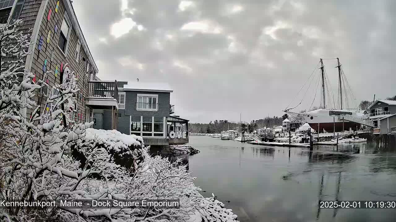A snow-covered waterfront scene shows buildings, a large sailboat docked, and a calm waterway under a cloudy sky.