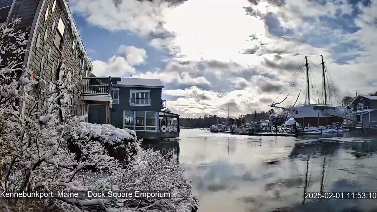 Snow-covered buildings and trees line a waterway where a large sailboat is docked under a partly cloudy sky.