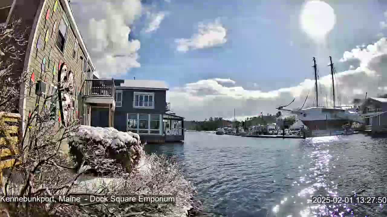 A partly sunny day with snow on the ground shows a waterfront view of colorful buildings, a large sailboat, and a calm waterway.