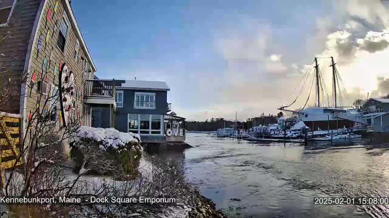 A snow-covered waterfront scene shows a building with colorful decorations next to a harbor where boats are docked under a partly cloudy sky.