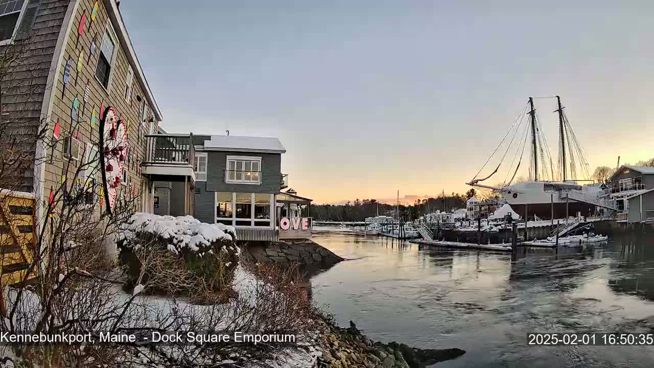 A snow-covered waterfront scene at sunset shows a building with colorful exterior art, another building with large windows, and a docked sailboat, all next to a calm river.