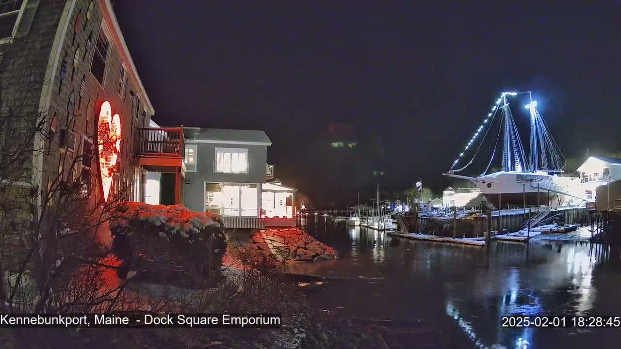 A snow-dusted waterfront scene at night features a building illuminated with a large red heart, other buildings with lit windows, and a large sailboat docked nearby.