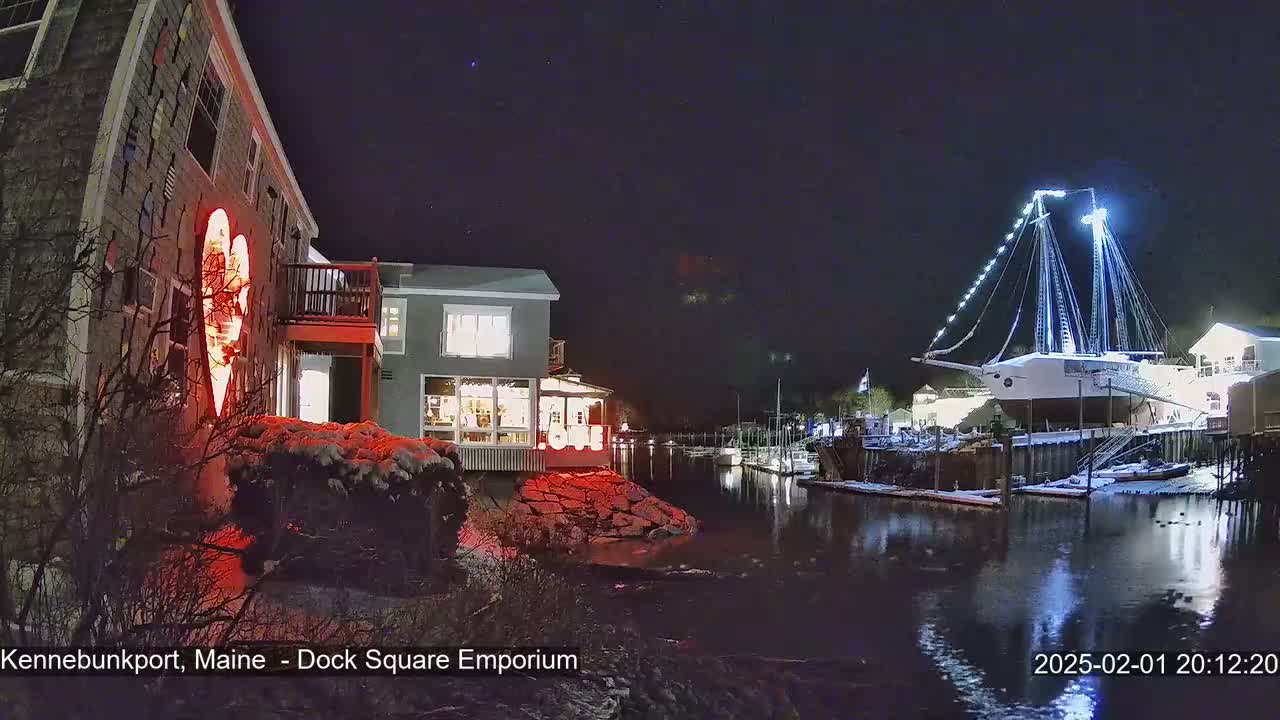 A nighttime scene shows a snow-dusted waterfront with a large, illuminated sailboat docked near buildings decorated with lights, one of which displays a large red heart.