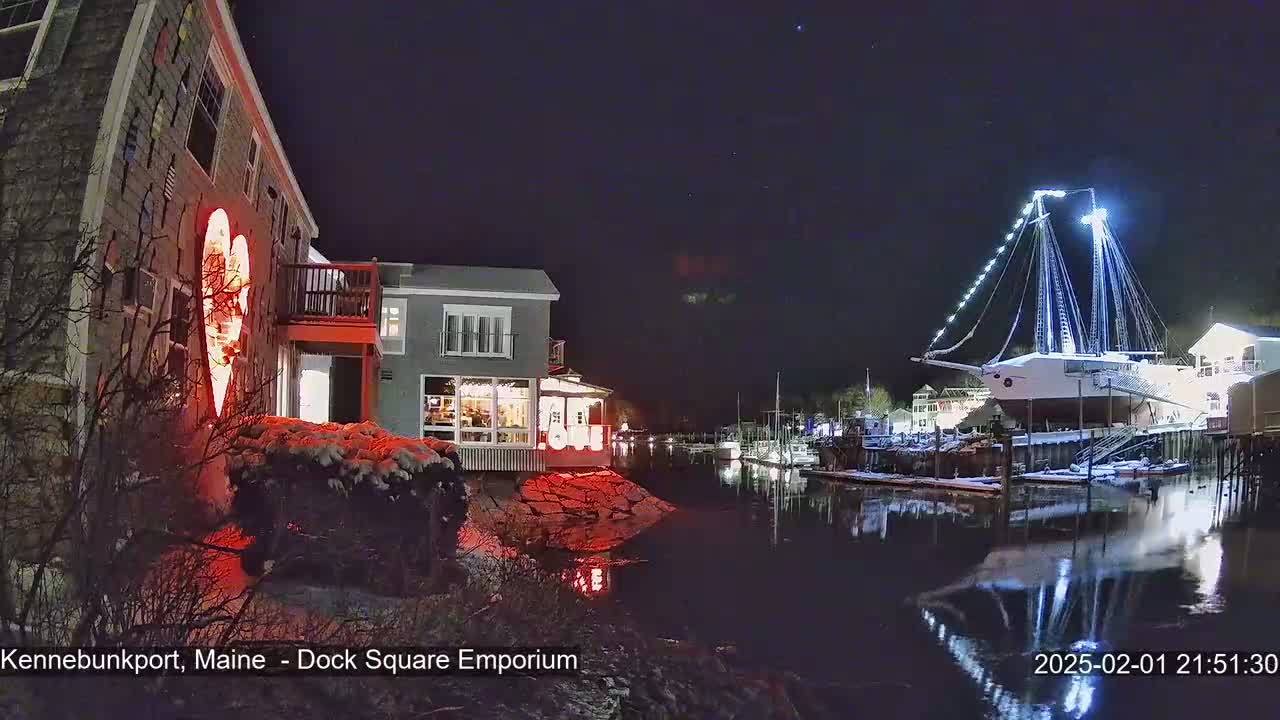A nighttime scene of a snow-dusted waterfront, featuring a building illuminated with a large red heart-shaped light display and a docked sailboat lit with white lights, reflecting on calm water under a dark, starry sky.