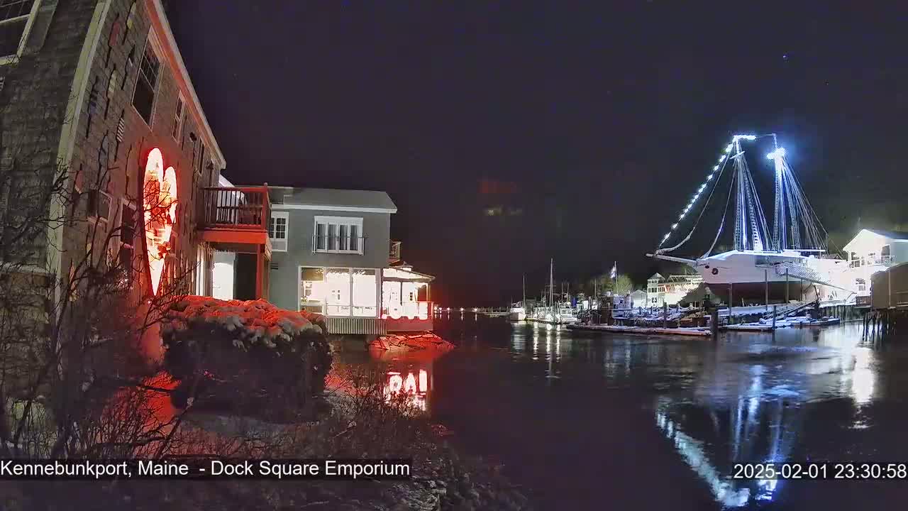A nighttime scene shows a snow-covered waterfront with buildings illuminated, a large sailboat docked, and a red heart-shaped light on the side of a building.