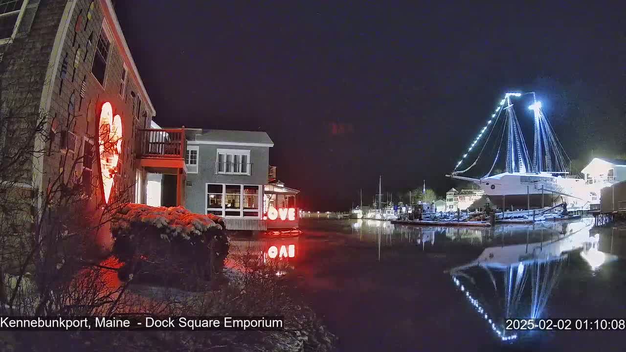 A snow-covered waterfront scene at night shows a building with a large illuminated heart, other buildings, and a large ship illuminated with lights, all reflected in calm water.