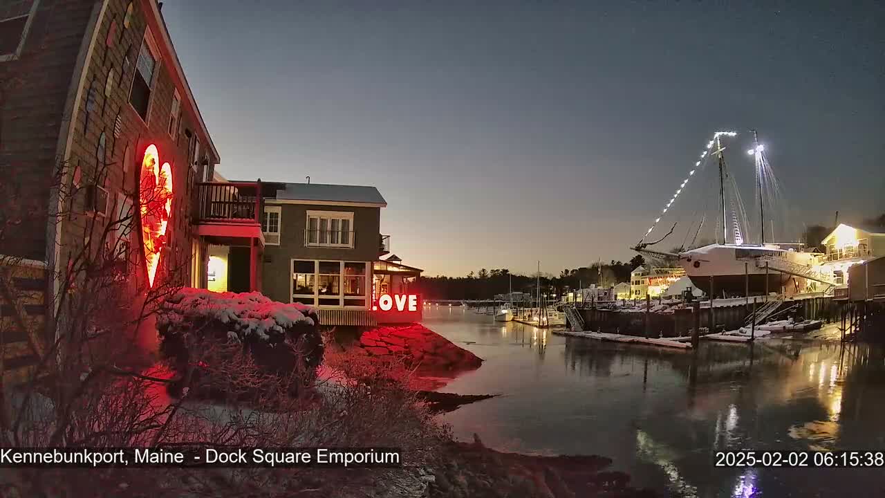 A snow-covered waterfront scene at twilight shows a building with a large illuminated heart and the word "LOVE" next to it, and a docked sailboat illuminated with lights.