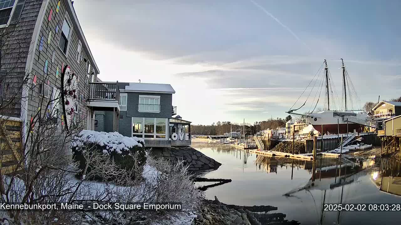 A snow-covered waterfront scene shows a harbor with a docked sailboat and buildings, under a partly cloudy sky.