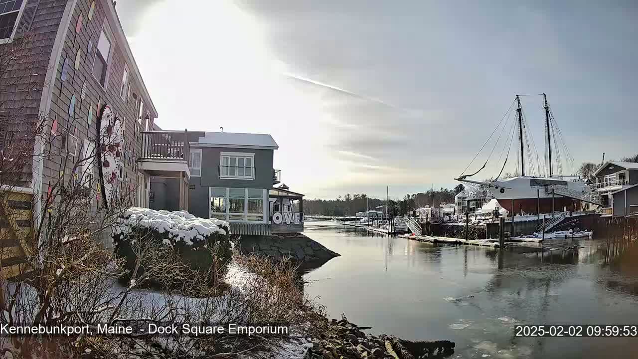 A snow-covered waterfront scene shows a calm waterway with a docked sailing ship, several buildings, and partially frozen water under a partly sunny sky.