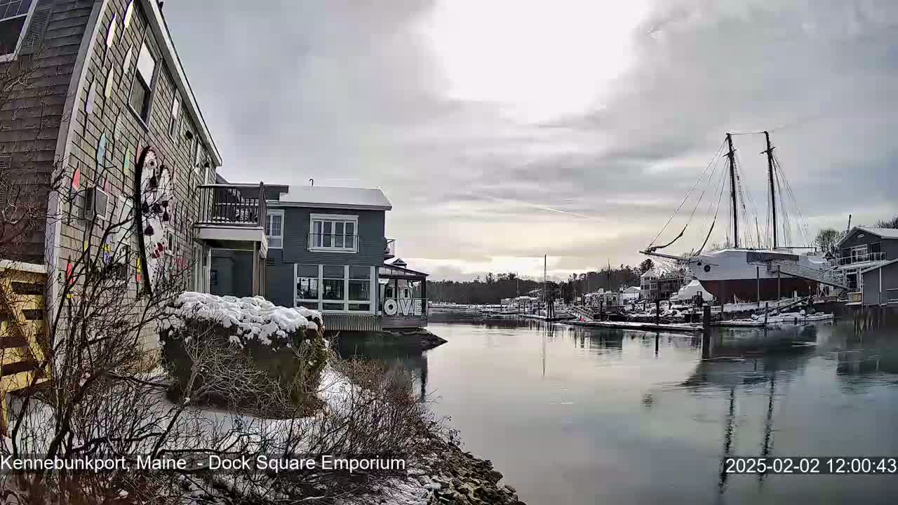 A snow-dusted waterfront scene shows a calm harbor with boats and buildings under a cloudy sky.