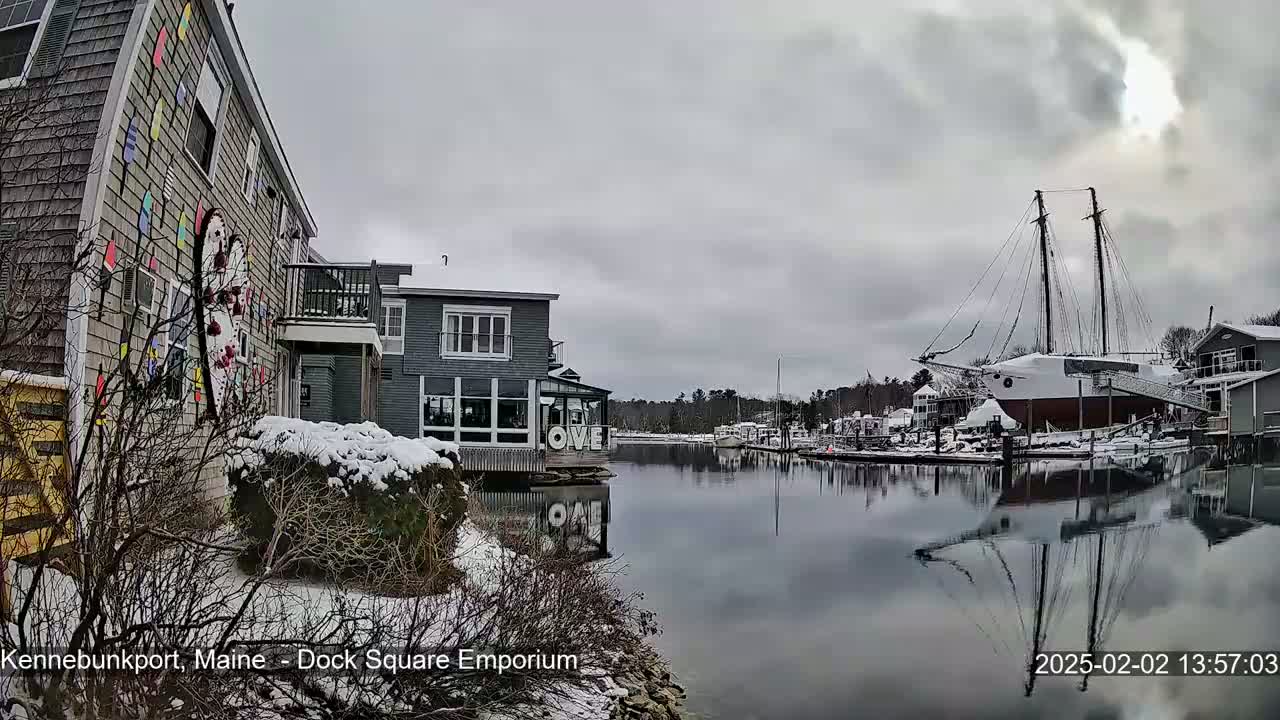 A snow-covered waterfront scene shows buildings, a large sailboat, and calm water reflecting the overcast sky.