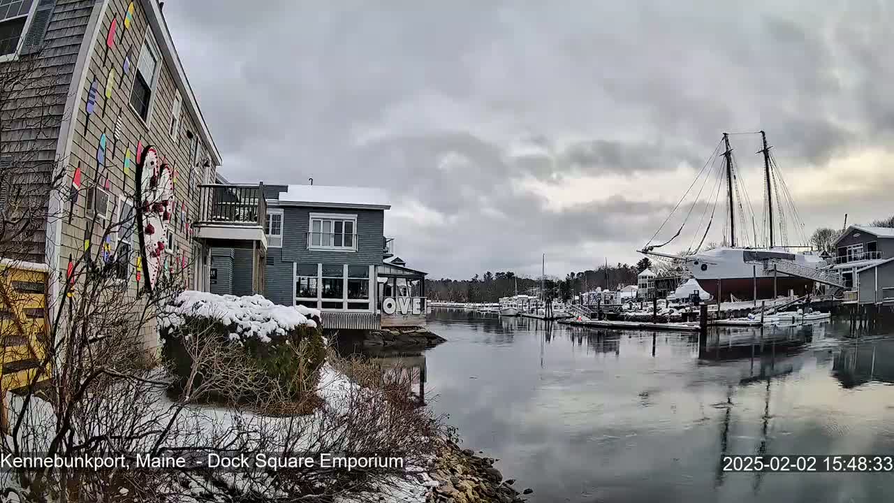 A snow-covered waterfront scene shows buildings, a large sailboat docked, and a calm waterway under an overcast sky.
