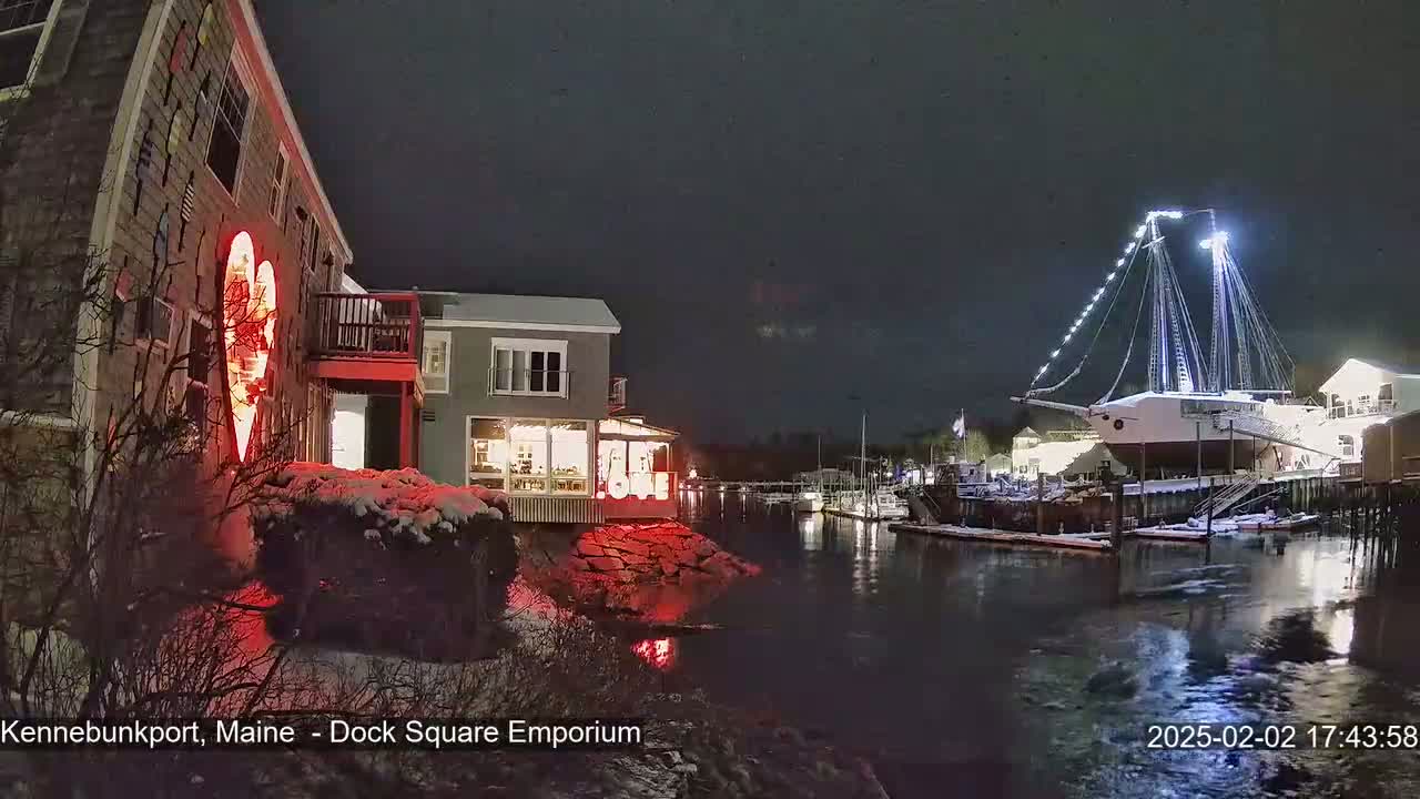 A snow-dusted waterfront scene at night shows a building with a large illuminated heart on its exterior, a docked sailboat lit up with lights, and other buildings and boats along a calm waterway.