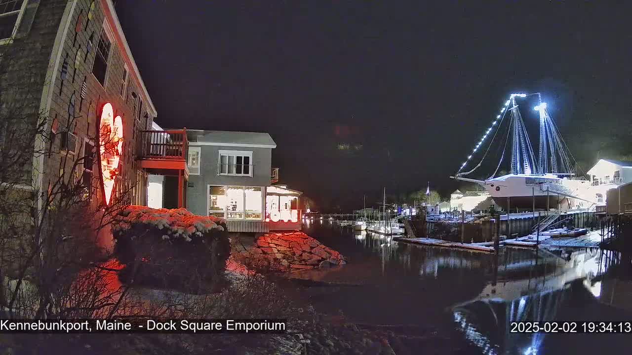 A snow-dusted waterfront scene at night shows a building with a large illuminated heart on its side, other buildings, a docked sailboat with lights, and calm water reflecting the lights.