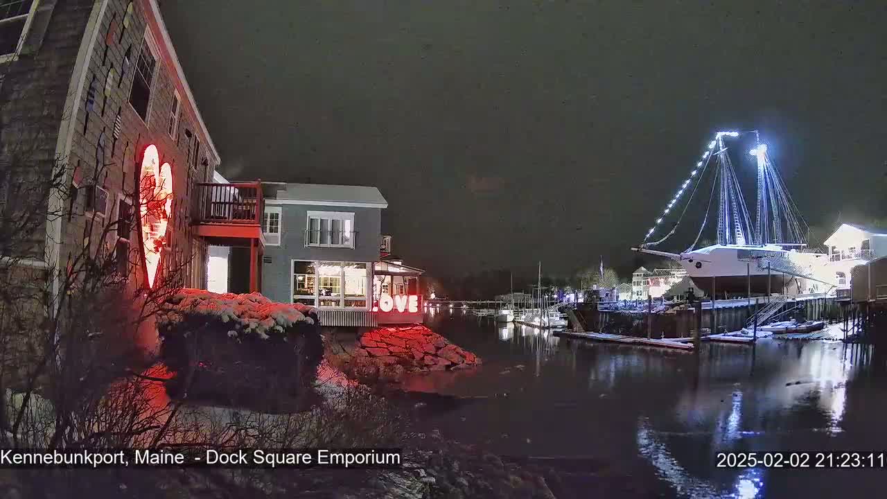 A nighttime scene of a snow-dusted waterfront shows a building with a large illuminated heart and the word "LOVE" displayed, and a large sailboat docked nearby under a dark, possibly snowy sky.