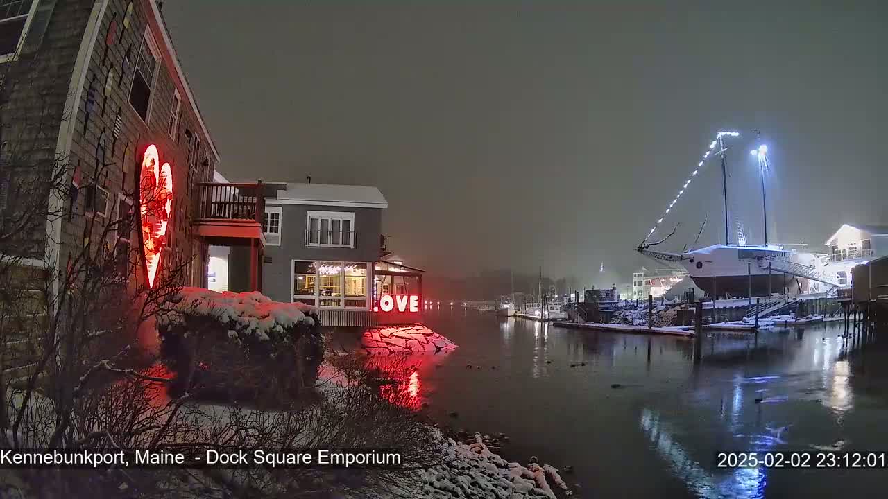 A snow-covered waterfront scene at night shows a building with a large illuminated heart and the word "LOVE" displayed, next to a docked sailboat illuminated with lights, all under a hazy, dark sky.