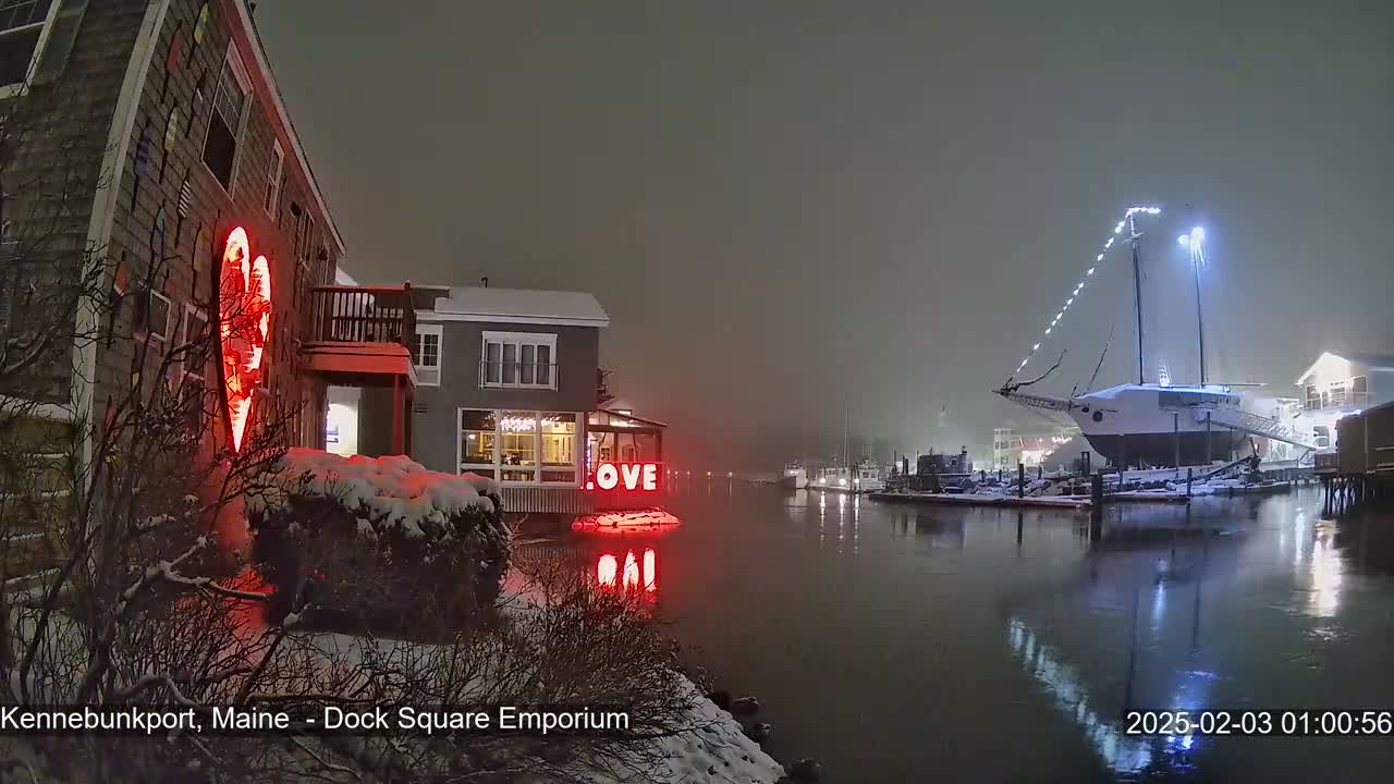 A snow-covered waterfront scene at night shows a building with a large illuminated heart and the word "LOVE" in lights, next to a snow-covered sailboat and other buildings.