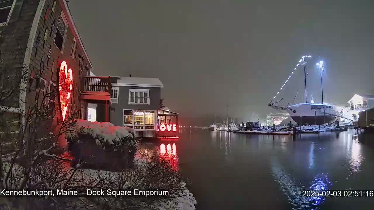 A snow-covered waterfront scene at night shows a building with a large illuminated heart, a lit-up "LOVE" sign, and a docked sailboat decorated with lights in the background.
