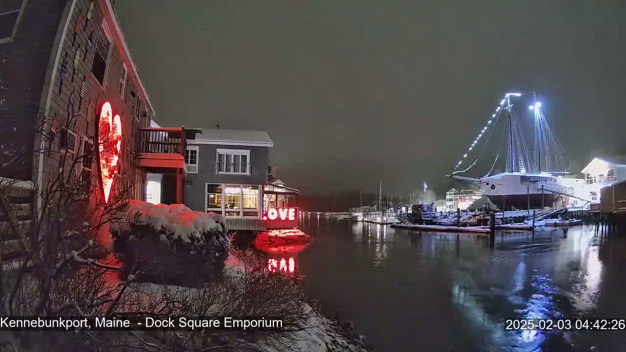 A snow-covered waterfront scene at night shows a building with a large illuminated heart, another building with the word "LOVE" in lights, and a docked sailboat, all illuminated under a dark, overcast sky.
