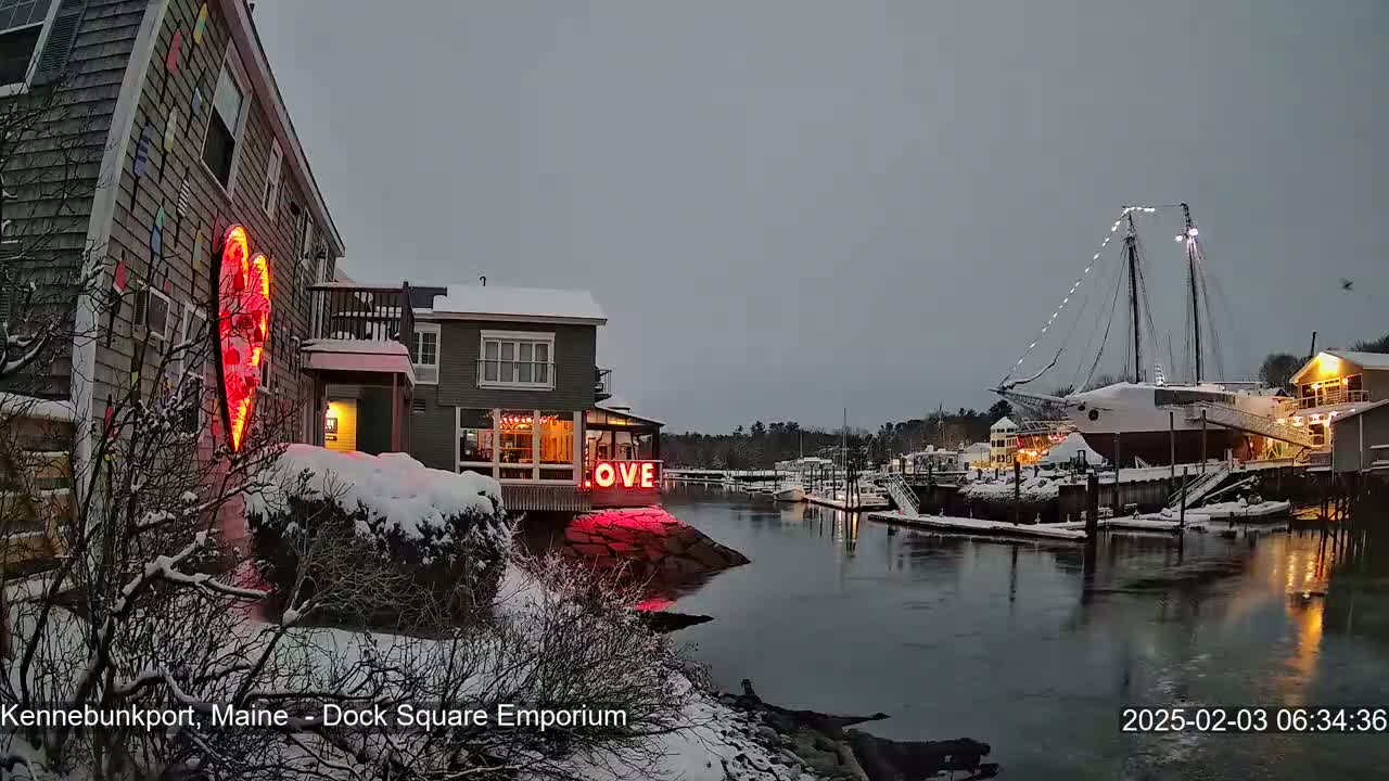 A snow-covered waterfront town at twilight features a building with a large illuminated heart and the word "LOVE" in lights, with a large sailing ship docked nearby.