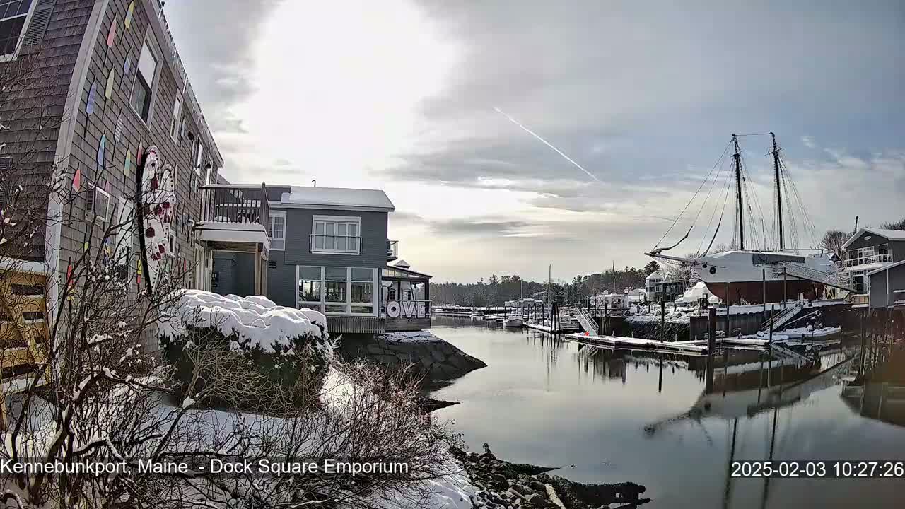 A snow-covered waterfront scene shows a canal with a large sailing ship docked, alongside buildings and trees dusted with snow under a partly cloudy sky.