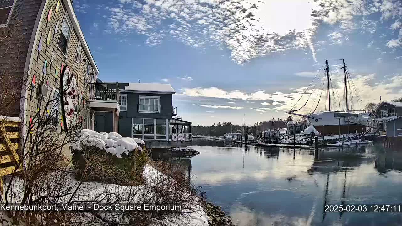 A snow-covered waterfront scene with colorful buildings, a large sailboat docked, and a partly cloudy blue sky.