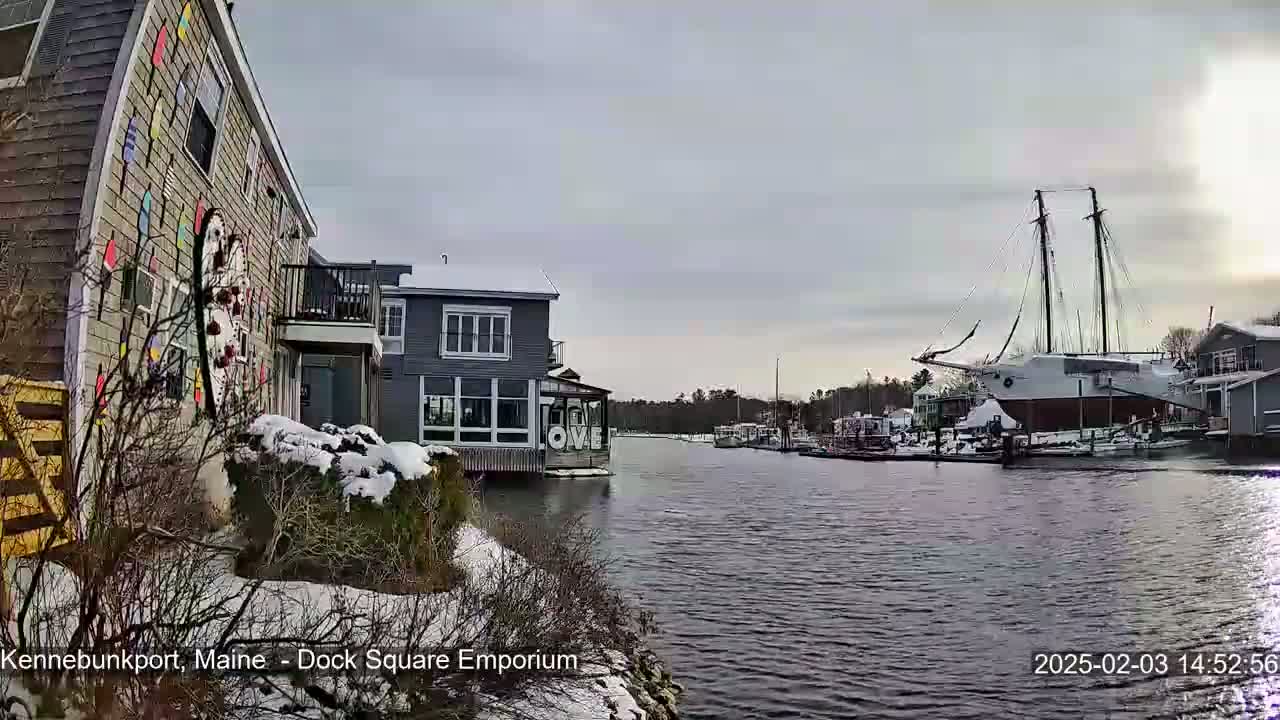 A snow-covered waterfront scene shows colorful buildings and a large sailboat docked in a calm waterway under an overcast sky.